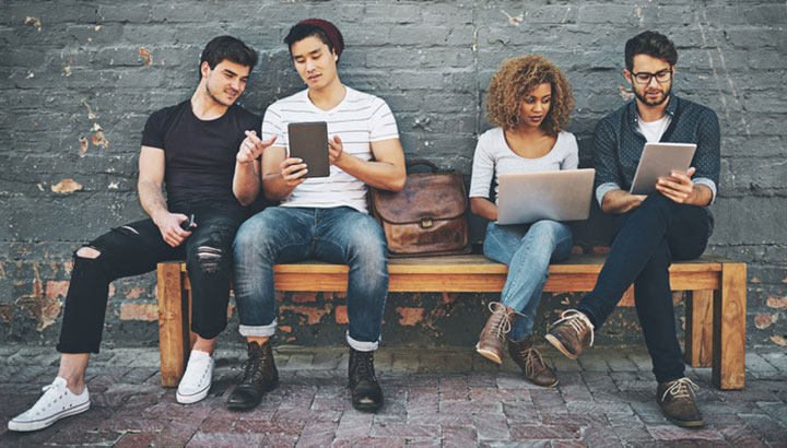 Four people sitting on a bench looking at laptops and iPads